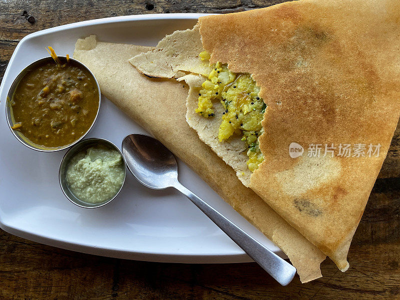 印度早餐masala dosa的图像，薄的折叠土豆煎饼放在白色盘子上，配上蘸料/调味品/酸辣酱，包括酸辣酱，高架俯视图